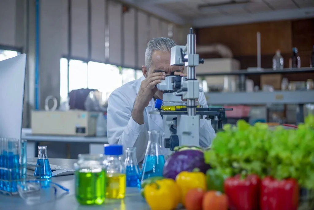A laboratory worker looking through a microscope beside fresh vegetables and glassware