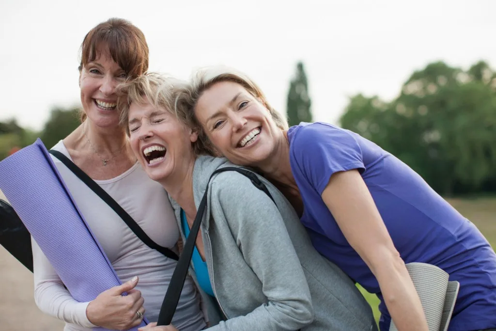 Three women smiling together outdoors with yoga mats