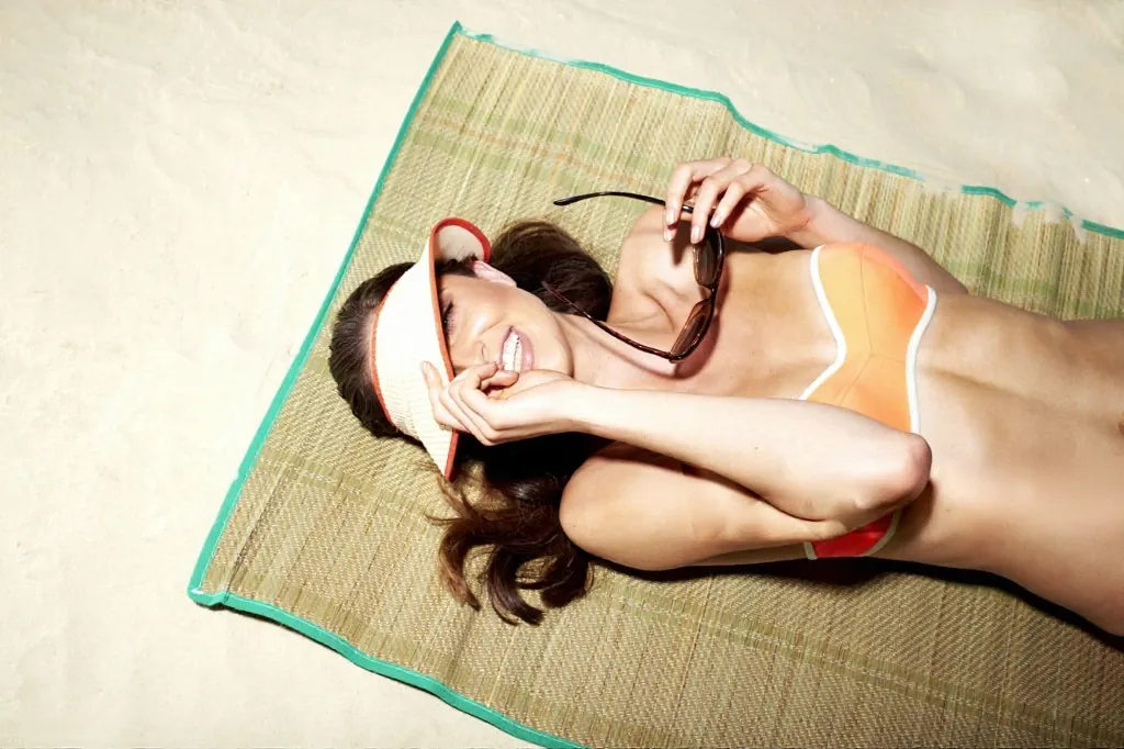 A woman relaxing in the summer sun on a woven mat, smiling with a visor shading her face